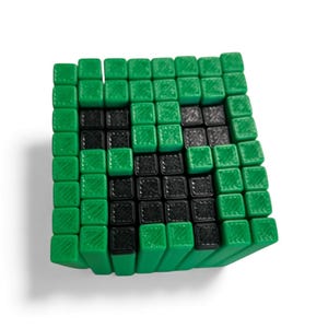 May include: A green and black pixelated cube, resembling a character from a video game. The cube is composed of numerous small, square blocks, with the green blocks forming the outer layer and the black blocks creating a face design. The cube is on a white background.