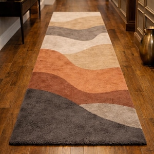Puede incluir: Una alfombra larga y rectangular con un diseño ondulado y en capas en tonos marrones, beige y grises. La alfombra está colocada sobre un suelo de madera, creando un ambiente cálido. El diseño de la alfombra se asemeja a un paisaje.
