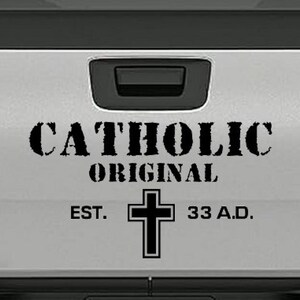 May include: A white truck tailgate with the words "CATHOLIC ORIGINAL" in black, above a black cross and the text "EST. 33 A.D."