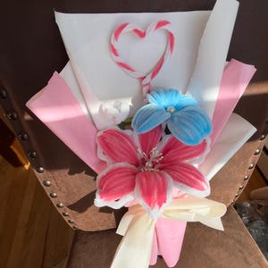 May include: A bouquet of handmade flowers in shades of pink and blue, wrapped in white and pink paper. The bouquet features a heart-shaped accent and a cream-colored ribbon. The flowers appear to be made of felt or a similar soft material.