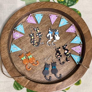 Peut inclure: Plusieurs paires de boucles d'oreilles en forme de chat sont présentées sur un plateau en bois. Les boucles d'oreilles sont de différentes couleurs, dont orange, noir et blanc, et présentent des nœuds. Une bannière avec les lettres "ROLIC" est suspendue.