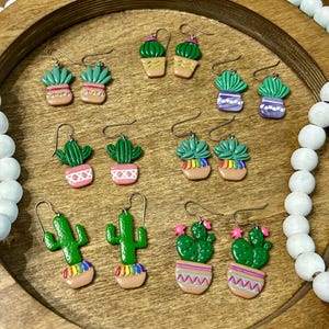 May include: A collection of six pairs of dangle earrings featuring miniature green cactus plants in various colorful pots. Each pair displays distinct patterns, including stripes, diamonds, and rainbow accents, suspended from silver-toned metal hooks.