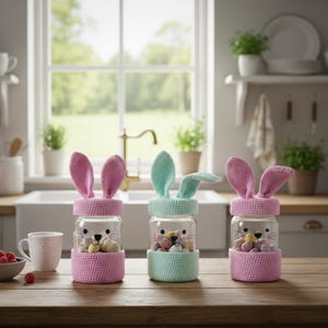 May include: Three glass jars decorated as Easter bunnies, each with a different colored crochet ear and base. The jars are filled with colorful candies and sit on a wooden table. The bunny faces are drawn on the glass.