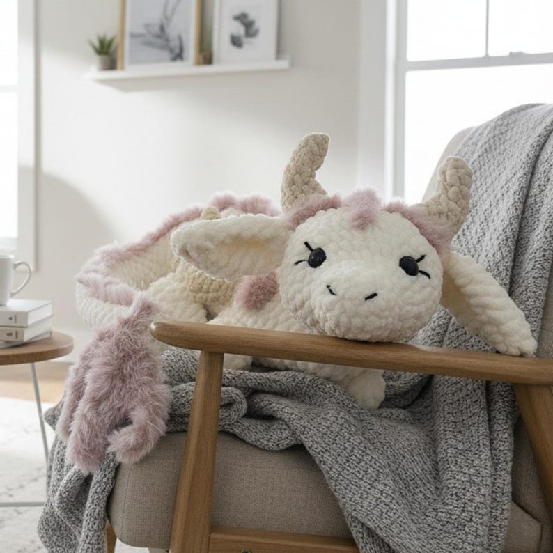 May include: A handmade, plush cow toy in cream and pink, with black eyes and a smiling face. The cow is lying on a gray blanket draped over a wooden chair. The toy has a soft, textured appearance, with fluffy accents.