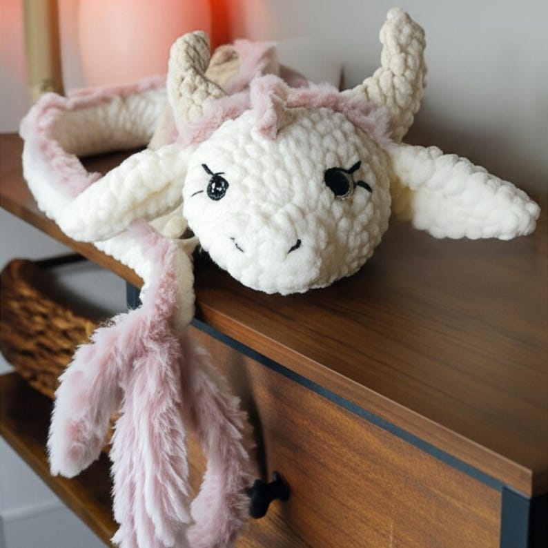 May include: A handmade, plush white dragon toy with pink accents. The dragon features a fluffy, textured body, large black eyes, and a long, pink, furry tail. The dragon is lying on a wooden surface, showcasing its soft, cuddly design.