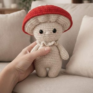 May include: A crocheted mushroom doll with a red cap and a beige body. The doll has black eyes and a ruffled collar. The mushroom is held in a hand against a neutral background.