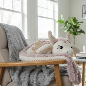 May include: A handmade, plush dragon pillow in white, beige, and pink. The dragon has large black eyes, small horns, and a fluffy pink mane. The pillow is resting on a wooden chair with a gray blanket.