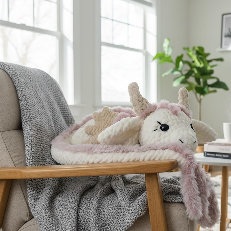 May include: A handmade, plush dragon pillow in white, beige, and pink. The dragon has large black eyes, small horns, and a fluffy pink mane. The pillow is resting on a wooden chair with a gray blanket.