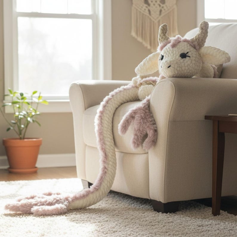 May include: A plush, cream-colored dragon with pink accents, including a long tail and limbs, rests on a cream armchair. The dragon has horns, wings, and a sweet face. The toy is designed to be soft and cuddly.