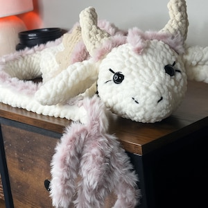 May include: A handmade, plush cow toy in white, cream, and pink. The cow features black button eyes, a stitched smile, and fluffy pink accents around the horns and body. The toy is made of soft, textured yarn.