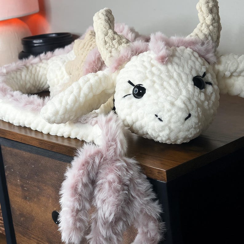 May include: A handmade, plush cow toy in white, cream, and pink. The cow features black button eyes, a stitched smile, and fluffy pink accents around the horns and body. The toy is made of soft, textured yarn.