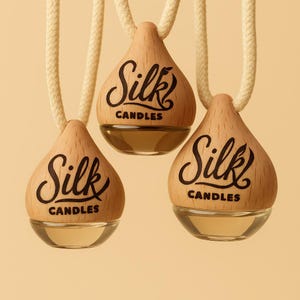 May include: Three teardrop-shaped air fresheners with wooden tops and glass bottoms, suspended by cream-coloured cords. The wooden tops are branded with the words "Silk Candles" in black script. The liquid inside is amber-coloured.