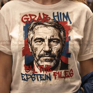 May include: Cream-colored t-shirt featuring a graphic portrait with the text "GRAB HIM BY THE EPSTEIN FILES." The portrait is overlaid with newspaper clippings and set against a red and blue backdrop. The text is in a graffiti-style font.