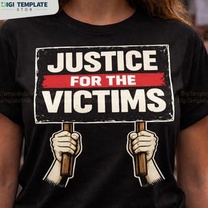 May include: Black t-shirt featuring a sign with the words "JUSTICE FOR THE VICTIMS" in white letters on a black background with a red stripe. The sign is held by two hands with wooden handles.