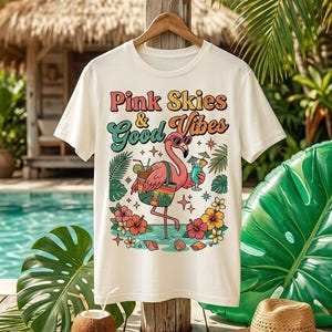Könnte beinhalten: Cremefarbenes T-Shirt mit einem Flamingo-Motiv, der eine Sonnenbrille trägt und einen Cocktail hält. Der Schriftzug "Pink Skies & Good Vibes" ist in bunten Retro-Lettern gehalten. Tropische Blumen und Palmenblätter umgeben den Flamingo.