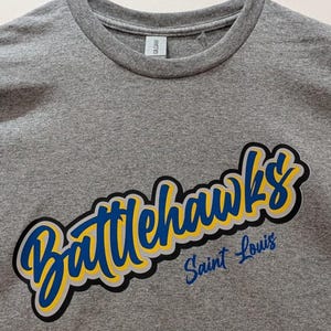 Puede incluir: Camiseta gris de cuello redondo con la palabra "Battlehawks" en fuente azul y amarilla, delineada en negro y gris. Debajo, "Saint Louis" est&aacute; escrito en azul m&aacute;s peque&ntilde;o.