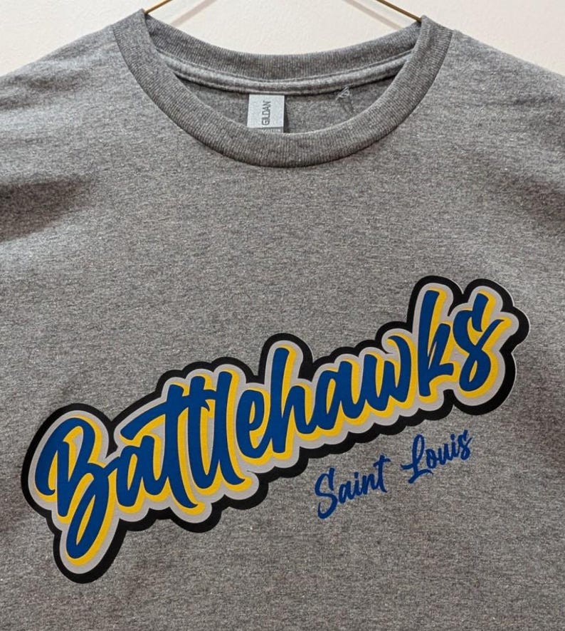 Puede incluir: Camiseta gris de cuello redondo con la palabra "Battlehawks" en fuente azul y amarilla, delineada en negro y gris. Debajo, "Saint Louis" est&aacute; escrito en azul m&aacute;s peque&ntilde;o.