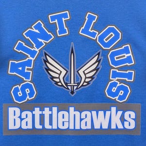 May include: A blue graphic design featuring the words "Saint Louis" in a curved, white-outlined font above the word "Battlehawks" in a rectangular banner. A sword and wing emblem is centered.