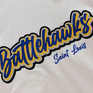 Puede incluir: Camiseta blanca con el texto "Battlehawks Saint Louis". La palabra "Battlehawks" es azul con un contorno amarillo y un borde negro. "Saint Louis" es azul, todo sobre un fondo blanco.