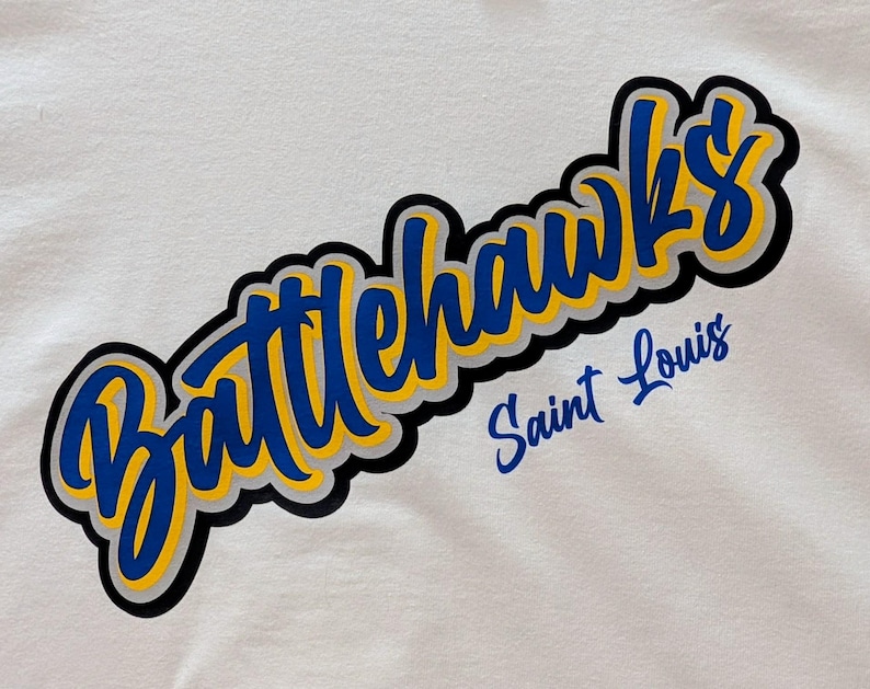 Puede incluir: Camiseta blanca con el texto "Battlehawks Saint Louis". La palabra "Battlehawks" es azul con un contorno amarillo y un borde negro. "Saint Louis" es azul, todo sobre un fondo blanco.