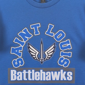 May include: A blue crew neck t-shirt with the words "Saint Louis" arched above a graphic of a sword with wings. Below the graphic is the word "Battlehawks" in white letters on a gray rectangle.