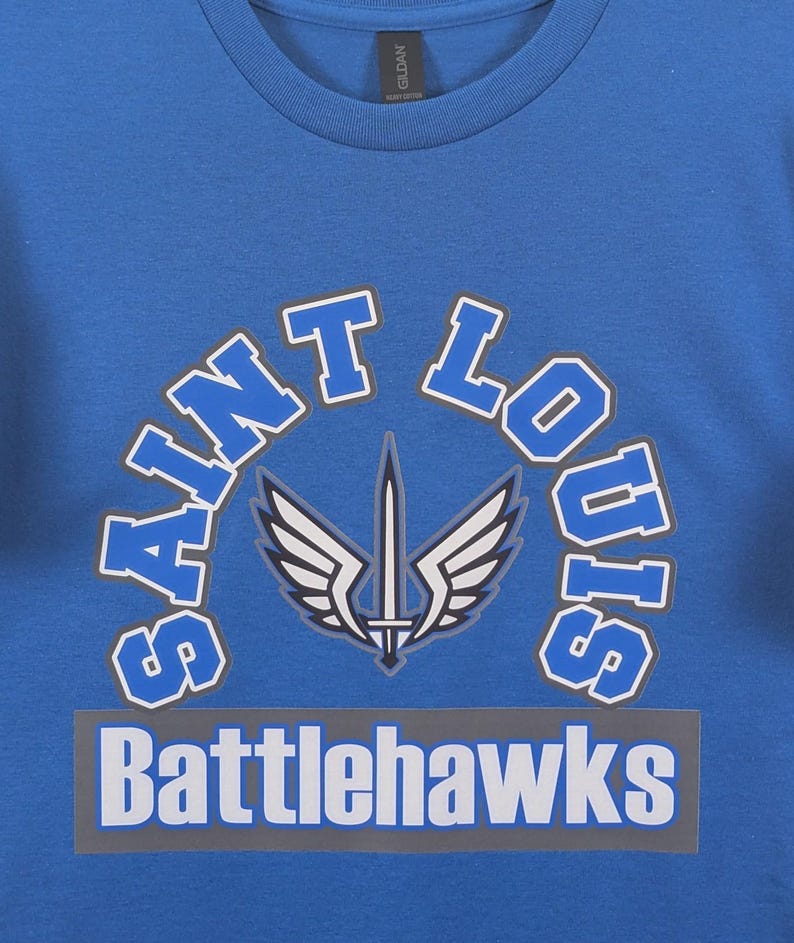 May include: A blue crew neck t-shirt with the words "Saint Louis" arched above a graphic of a sword with wings. Below the graphic is the word "Battlehawks" in white letters on a gray rectangle.