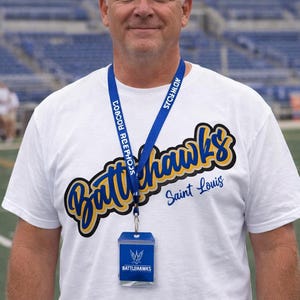Puede incluir: Una persona lleva una camiseta blanca con las palabras "Battlehawks Saint Louis" en letras azules y amarillas. Tambi&eacute;n lleva un cord&oacute;n azul con texto y un porta-credenciales transparente con un logotipo.