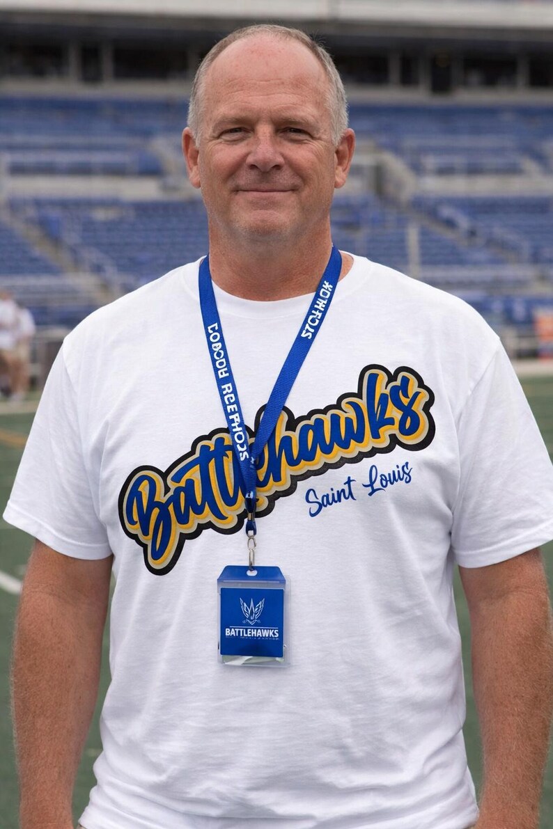 Puede incluir: Una persona lleva una camiseta blanca con las palabras "Battlehawks Saint Louis" en letras azules y amarillas. Tambi&eacute;n lleva un cord&oacute;n azul con texto y un porta-credenciales transparente con un logotipo.