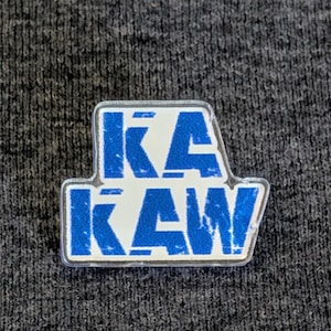 May include: A clear, rectangular pin with the word "KA KAW" in blue block letters. The pin has a white background and a blue border. The pin is on a gray, textured surface.
