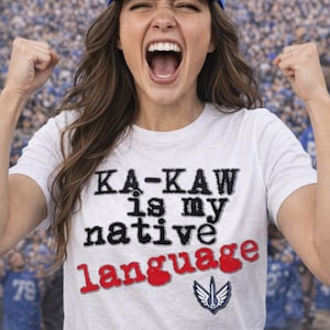 May include: White t-shirt with the text "KA-KAW is my native language" in black and red. A blue baseball cap with a white logo is worn. The background shows a crowd of people.