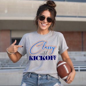 May include: A gray t-shirt with the text "Classy until Kickoff" in blue and orange. The person in the image is holding a brown football.