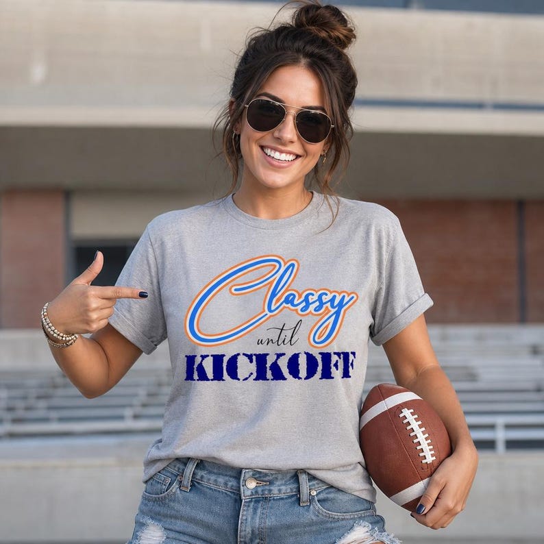 May include: A gray t-shirt with the text "Classy until Kickoff" in blue and orange. The person in the image is holding a brown football.