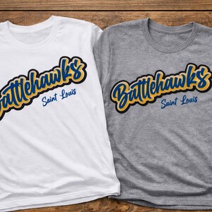 Puede incluir: Dos camisetas, una blanca y otra gris, cada una con el texto "Battlehawks Saint Louis" en una escritura estilizada. El texto es en azul y amarillo con un contorno negro. Las camisetas est&aacute;n sobre una superficie de madera.