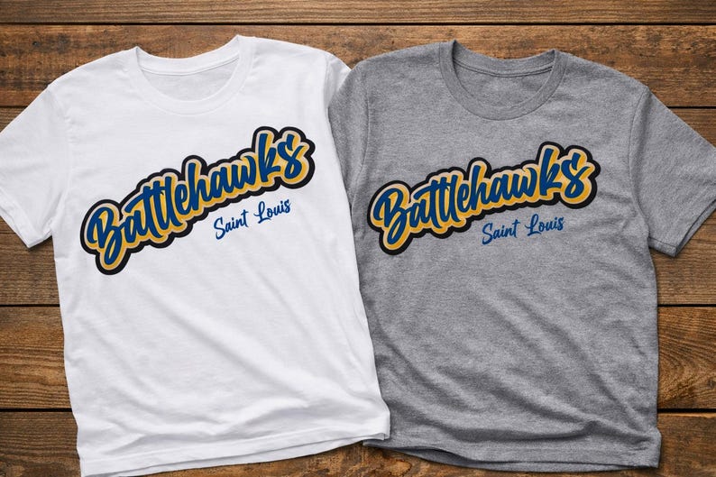 Puede incluir: Dos camisetas, una blanca y otra gris, cada una con el texto "Battlehawks Saint Louis" en una escritura estilizada. El texto es en azul y amarillo con un contorno negro. Las camisetas est&aacute;n sobre una superficie de madera.