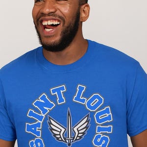 May include: Royal blue t-shirt with the words "Saint Louis Battlehawks" in white and blue lettering. The shirt features a logo of a sword and wings. The person wearing the shirt is smiling.