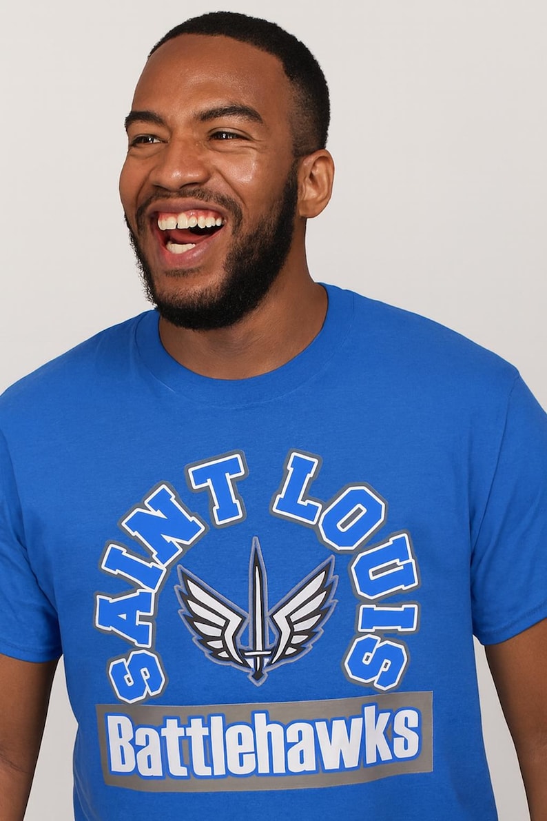 May include: Royal blue t-shirt with the words "Saint Louis Battlehawks" in white and blue lettering. The shirt features a logo of a sword and wings. The person wearing the shirt is smiling.