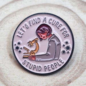 Peut inclure: Un pin's rond en émail sur fond rose clair représentant une personne en costume gris penchée sur un microscope doré. Le texte "LET'S FIND A CURE FOR STUPID PEOPLE" est inscrit en noir sur le pourtour, avec de petites icônes de virus noires.