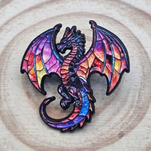 May include: A metal enamel pin shaped like a dragon with wings spread. The dragon features a black outline and a colourful stained glass effect on its body and wings, displaying vibrant shades of purple, pink, orange, and blue in a galaxy pattern.