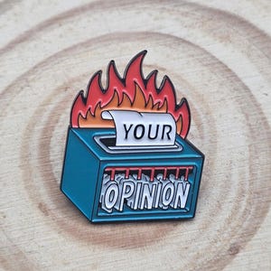 Peut inclure: Une broche en émail bleu sarcelle en forme de déchiqueteuse à papier en feu. La fente supérieure présente une bande de papier blanche avec le texte noir YOUR, tandis que l'avant affiche le texte blanc OPINION sur des bandes déchiquetées.