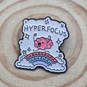 Peut inclure: Une broche en émail blanc représentant un cerveau rose de dessin animé avec une expression joyeuse. Le texte HYPERFOCUS est imprimé au-dessus du cerveau. En dessous, un arc-en-ciel coloré est entouré de nuages avec le texte AHAHAHAHAHAHAHAH.