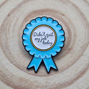 May include: A circular blue enamel pin shaped like a ribbon rosette with two tails. The centre is white with a gold border and features the black text "Didn't quit my job today" written in a cursive script. The pin has a black metal outline.