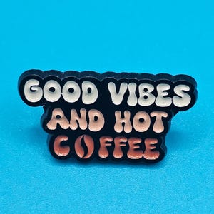 Peut inclure: Une broche en émail noir avec une police de style rétro ondulée. Le texte indique GOOD VIBES en blanc, suivi de AND HOT en rose clair, et COFFEE en rose clair avec une icône de grain de café remplaçant la lettre O. Bordure noire.