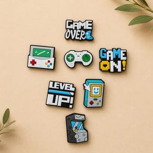 May include: A collection of seven colourful rubber shoe charms featuring video game themes. The designs include text reading GAME OVER, GAME ON, and LEVEL UP, alongside icons of a handheld console, a game controller, and an arcade machine.