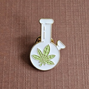 May include: A white enamel pin shaped like a water pipe or bong with gold-toned metal edges. The centre of the round base features a light green cannabis leaf graphic. The pin has a vertical neck and a small side attachment, finished with a gold backing.