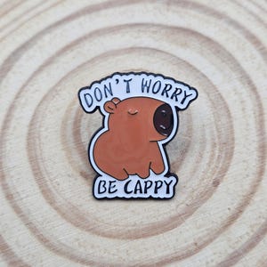 May include: A brown enamel pin shaped like a capybara with a dark brown nose and closed eyes. The pin features a white border with black text that reads DON'T WORRY BE CAPPY, with the top text arched over the head and the bottom text below.