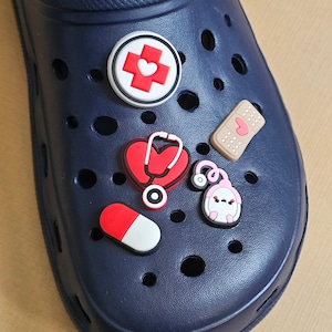 May include: A dark blue clog shoe decorated with five medical-themed charms. The charms include a white circle with a red cross and heart, a red heart-shaped stethoscope, a beige adhesive bandage, a red and white pill, and a pink medical device.