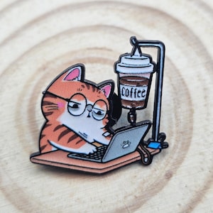 May include: An enamel pin featuring an orange tabby cat with glasses typing on a laptop. A black stand holds a coffee cup IV drip bag above the desk. The cup has the word COFFEE printed on it. The cat has pink ears and a white chest and face.
