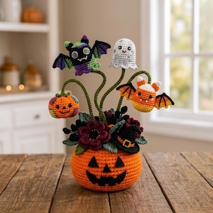 Può includere: Un centrotavola di Halloween all'uncinetto con una base a forma di zucca arancione, riempita di fiori viola scuro, foglie verdi e un cappello da strega nero. Quattro steli si innalzano, sormontati da una piccola zucca, un pipistrello viola, un fantasma bianco e un pipistrello a forma di caramella.