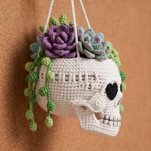 May include: A white crocheted skull planter hanging by white strings, filled with various crocheted succulents in shades of purple, light blue, and green. The skull features black heart-shaped eye sockets and a stitched mouth, with green vines trailing down.