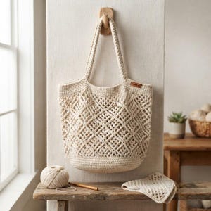Puede incluir: Un bolso de ganchillo de color crema con un patrón de malla en forma de diamante en el cuerpo, una base sólida y dos asas resistentes. Una pequeña etiqueta rectangular marrón está unida al lado superior derecho del bolso que cuelga de un gancho.
